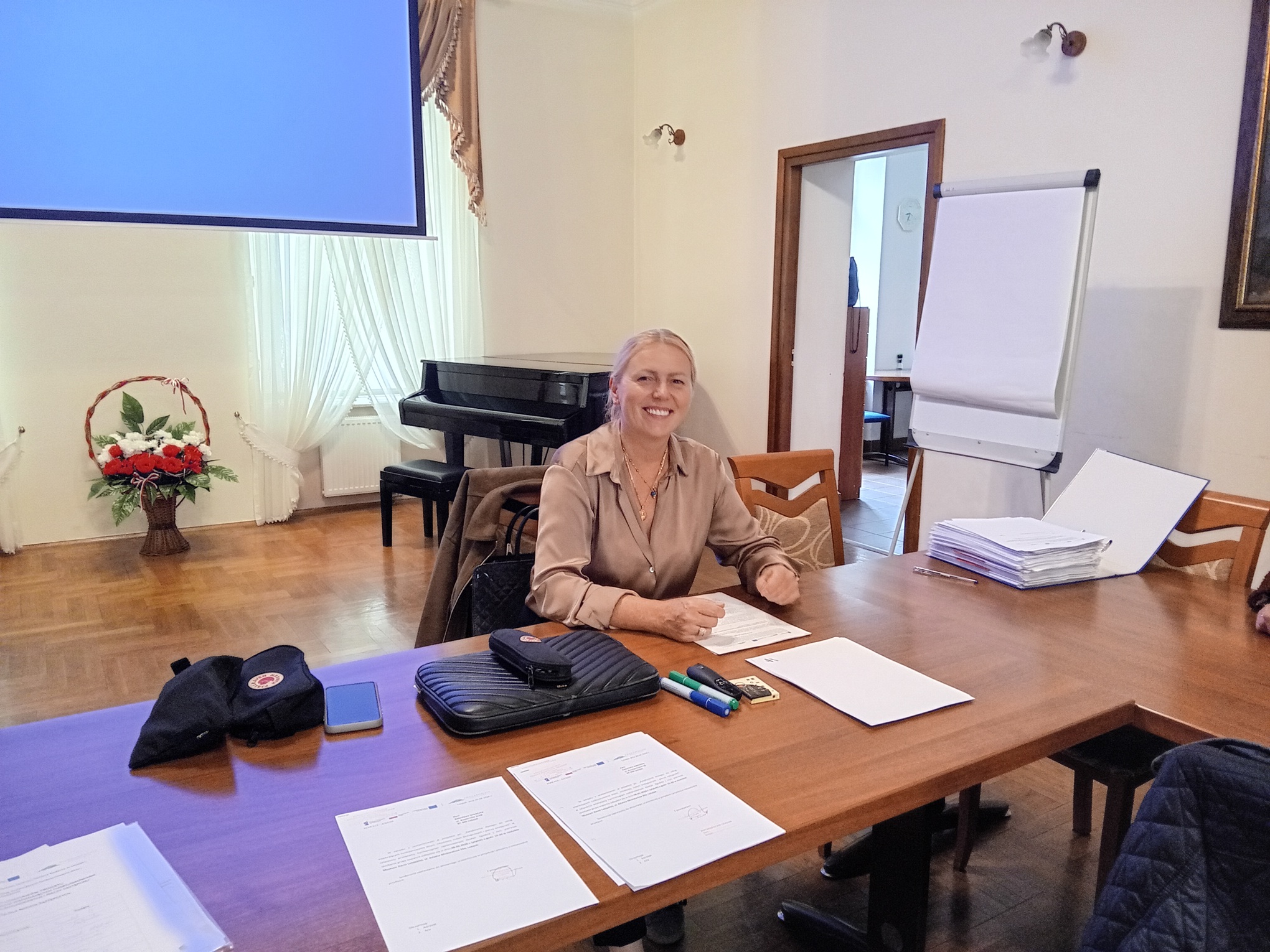Uśmiechnięta kobieta siedząca przy stole z&nbsp;dokumentami w&nbsp;sali konferencyjnej. W&nbsp;tle widać ekran projekcyjny, fortepian i&nbsp;kwiaty.